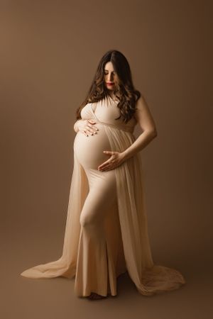 Elegant maternity portrait in beige tones showing woman in flowing dress cradling her belly against neutral backdrop.