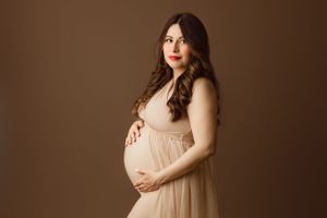 A pregnant woman in a flowing beige dress poses elegantly against a brown backdrop in a maternity photo.