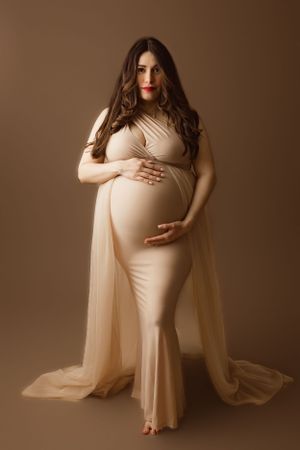 Elegant maternity portrait in beige dress with flowing cape against brown backdrop in studio setting.