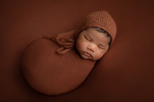 Newborn baby boy wearing a bonnet and wrapped, laying on his side, photographed by Pamela Clark