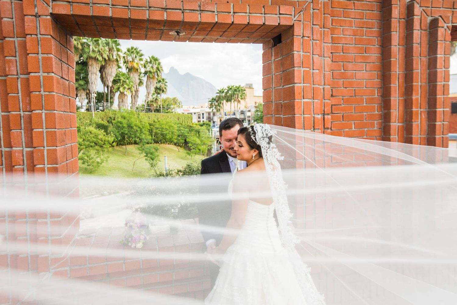 A flowing white veil catches the breeze through a brick archway during a romantic wedding photo shoot.