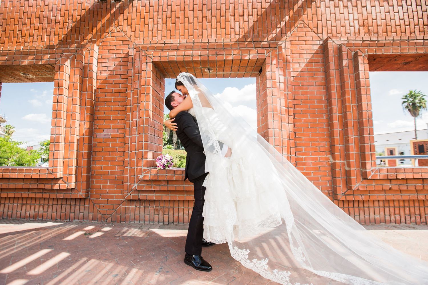 A playful wedding portrait framed by red brick architecture with dramatic lighting and flowing veil.