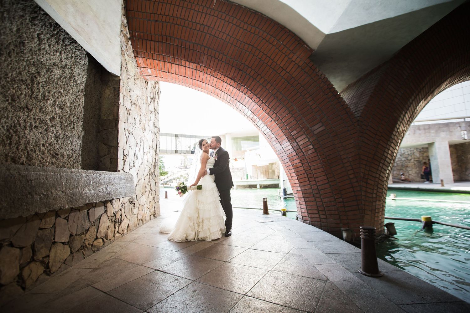 A romantic moment captured under a brick archway beside a reflecting pool.