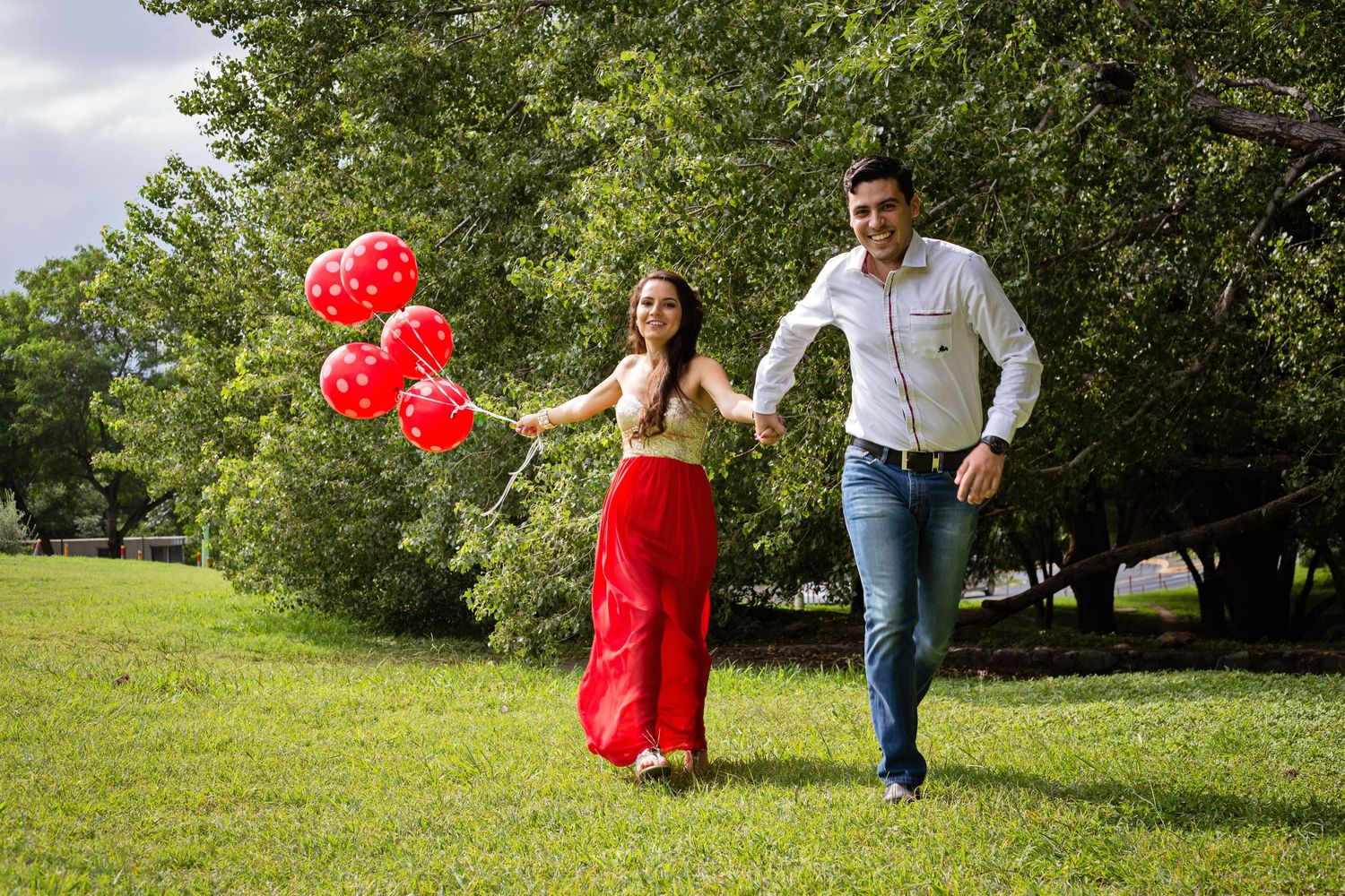 Couple runs through green park holding red balloons on sunny day.