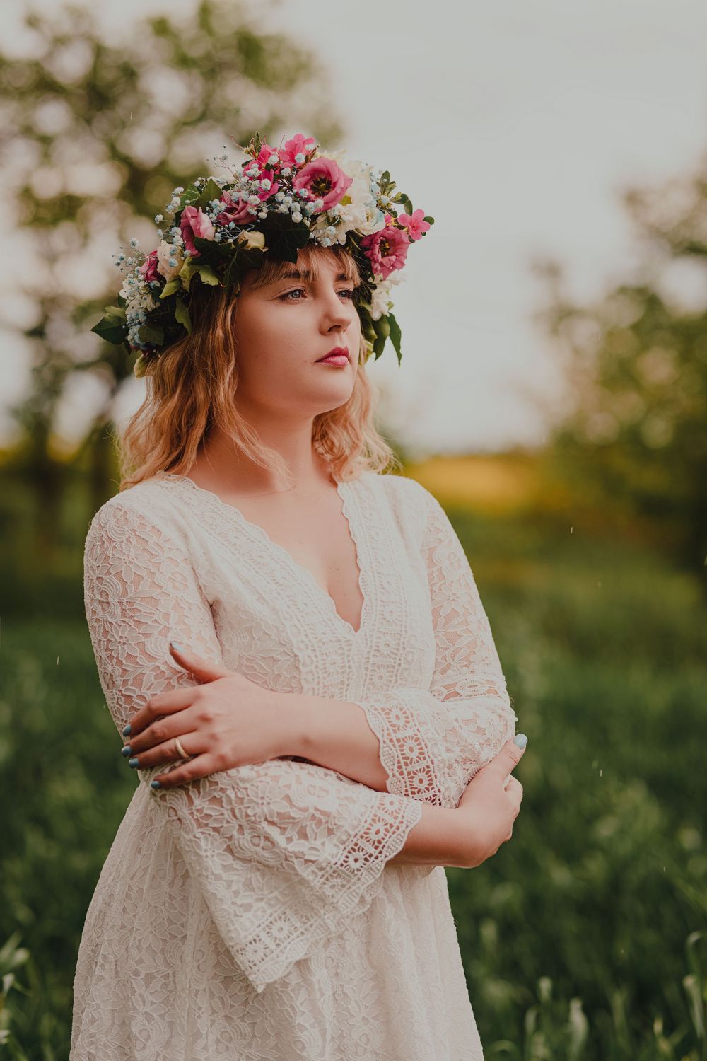 Figure in white lace dress with floral crown poses in a sunlit meadow at golden hour.