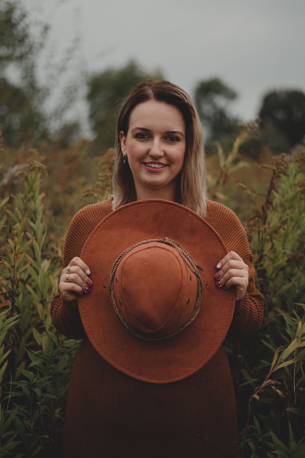 Person playfully posing with brown wide-brimmed hat in natural outdoor setting.