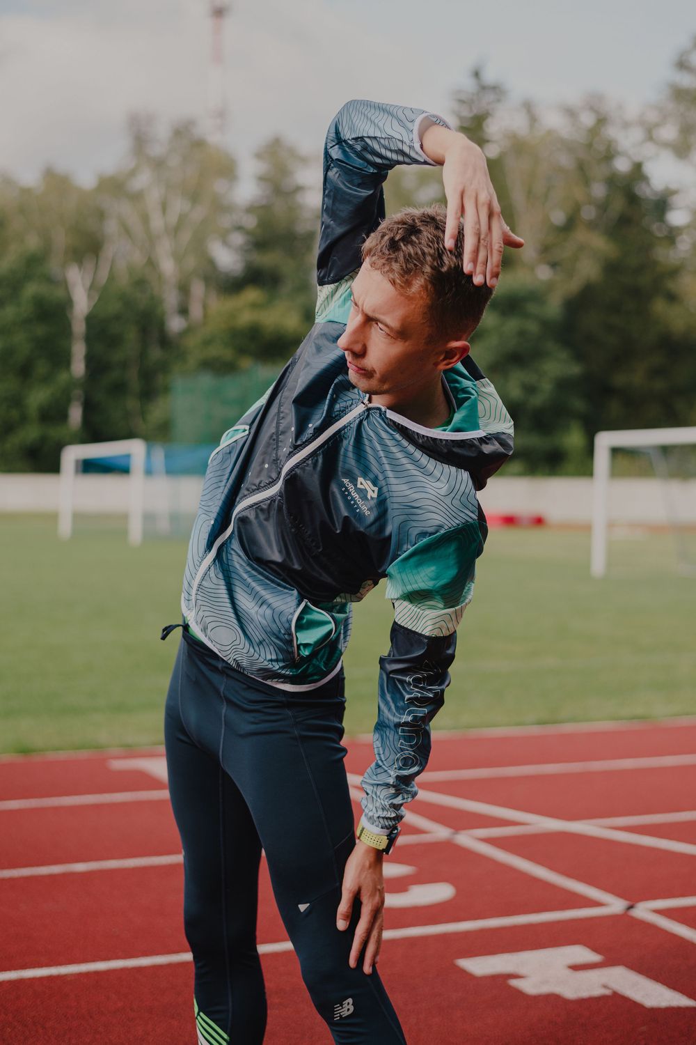 Athletic figure stretching in workout gear on a red running track with trees in background.