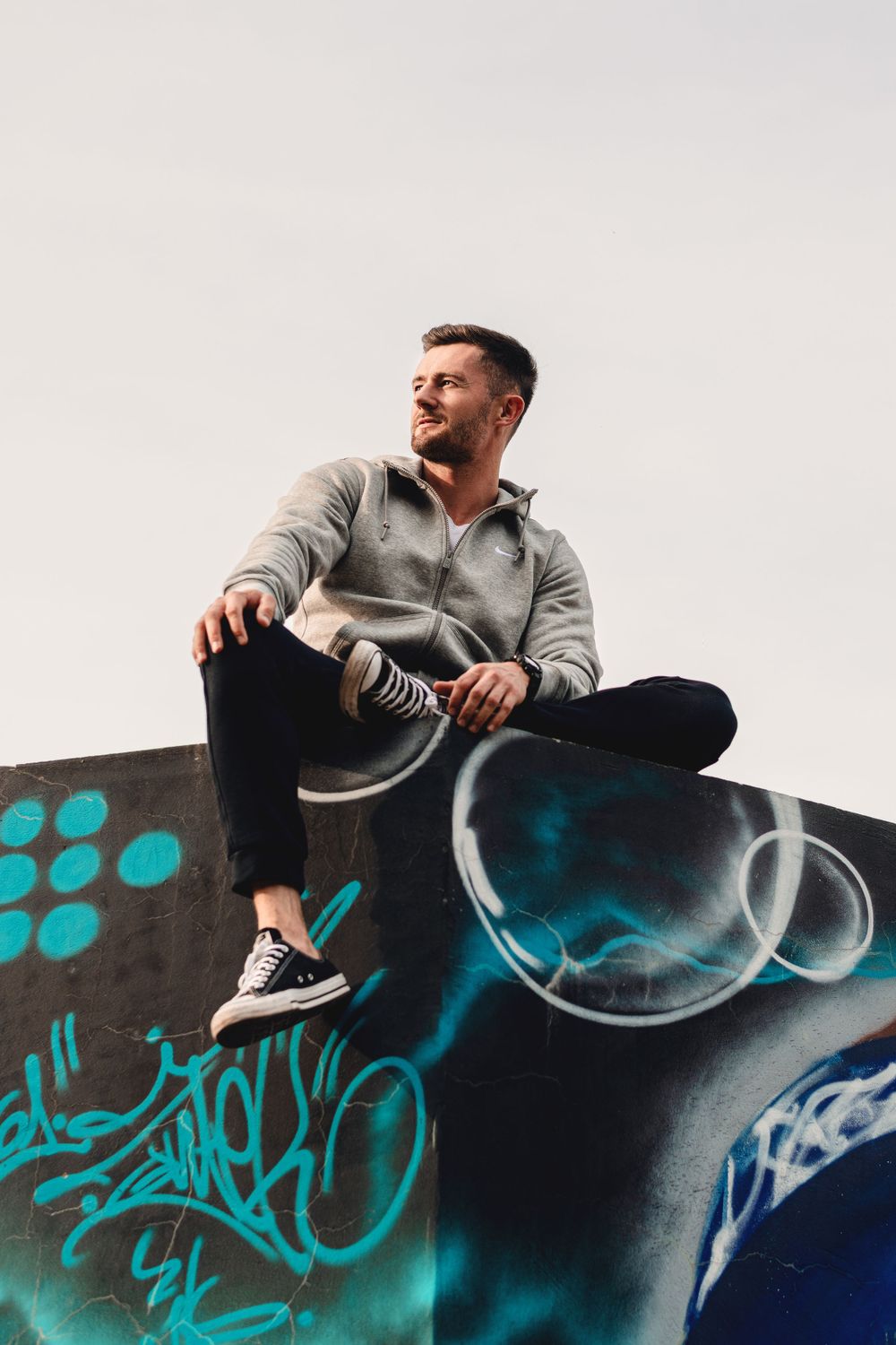 Person in grey hoodie sitting on graffiti-covered wall against bright sky.