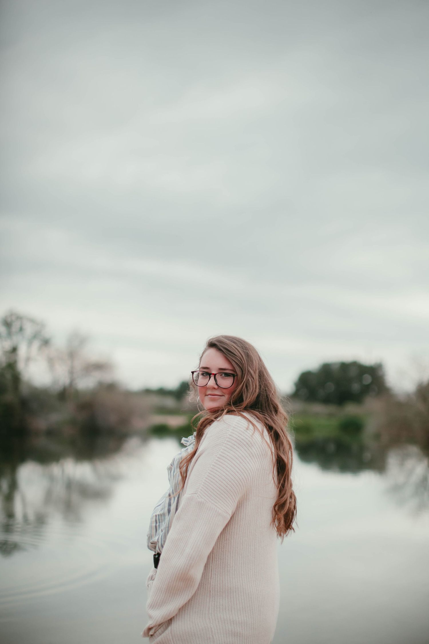 Person in cream sweater stands by misty lake during overcast day.