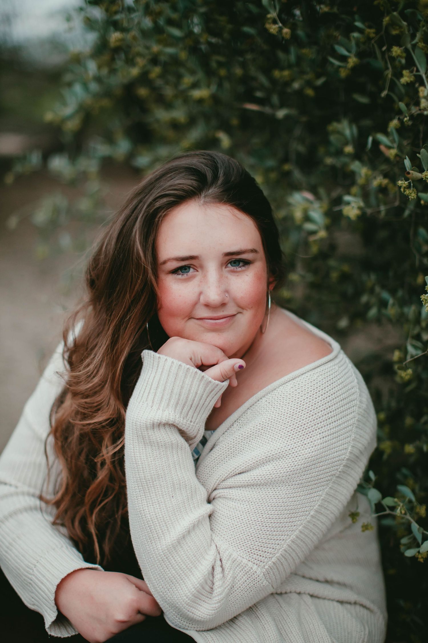 A cozy portrait in a cream sweater against green foliage in soft natural lighting.