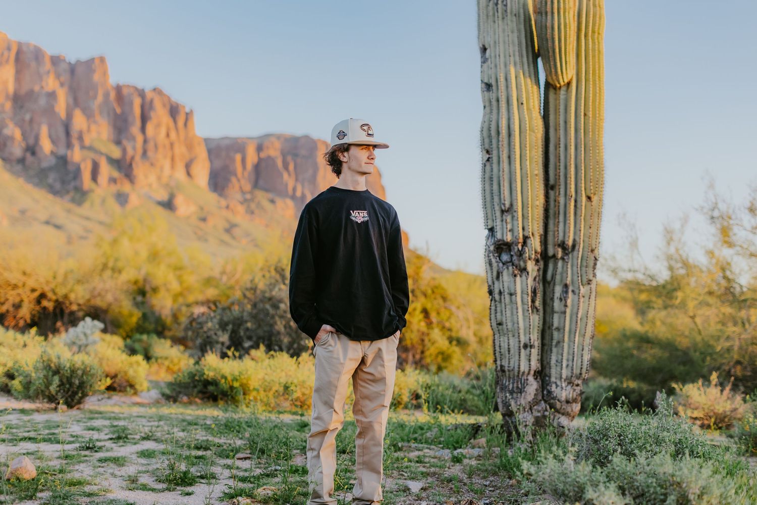 Person in casual wear standing next to tall saguaro cactus with Superstition Mountains in background.