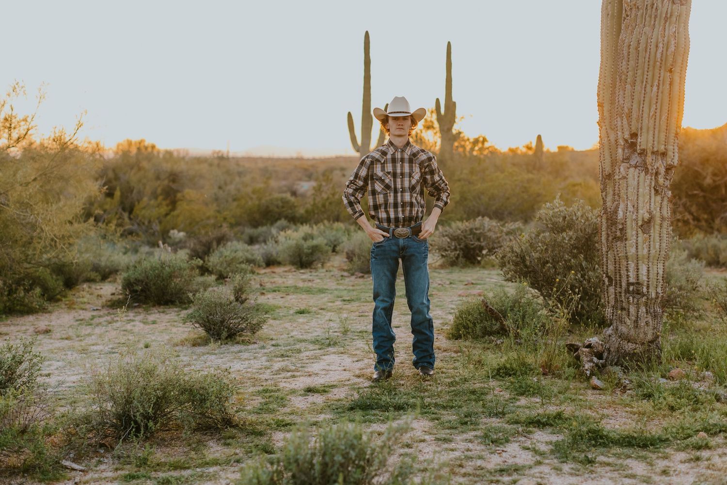 A person in western attire stands among desert cacti and brush during golden hour.