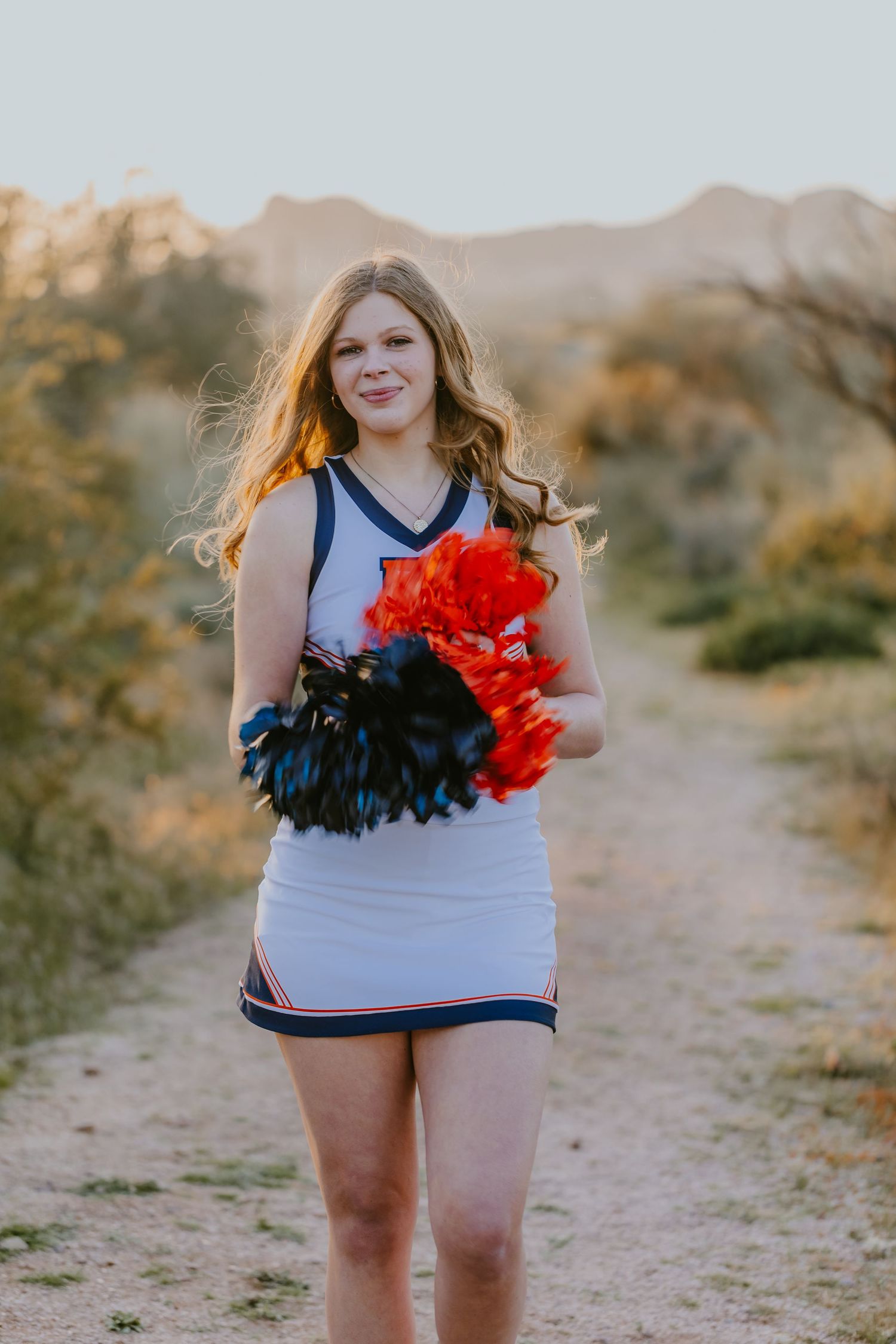 Cheerleader poses with red and black pom poms against desert landscape at sunset.