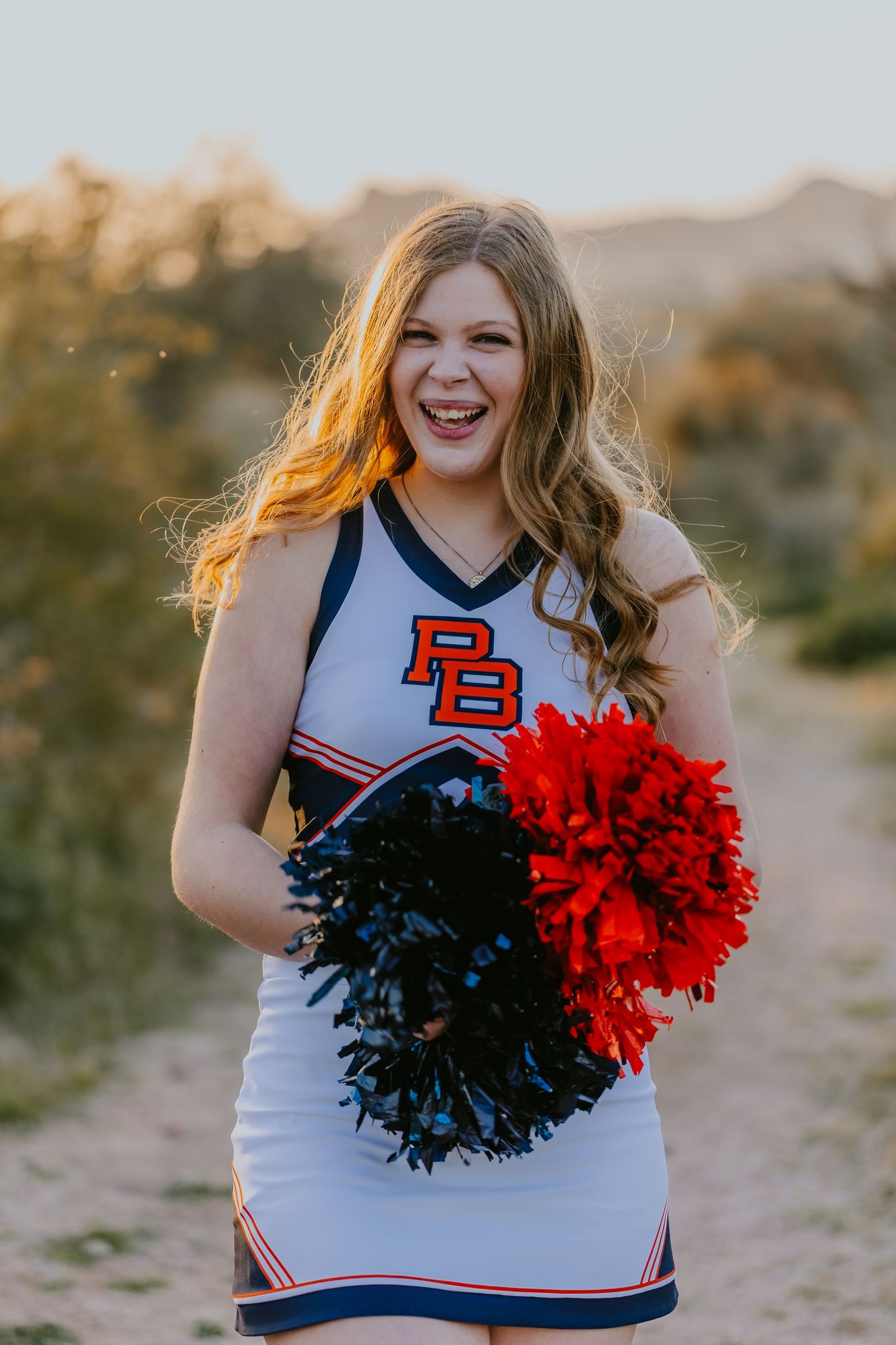 Cheerleader in white uniform holding red and black pom poms poses in desert setting.