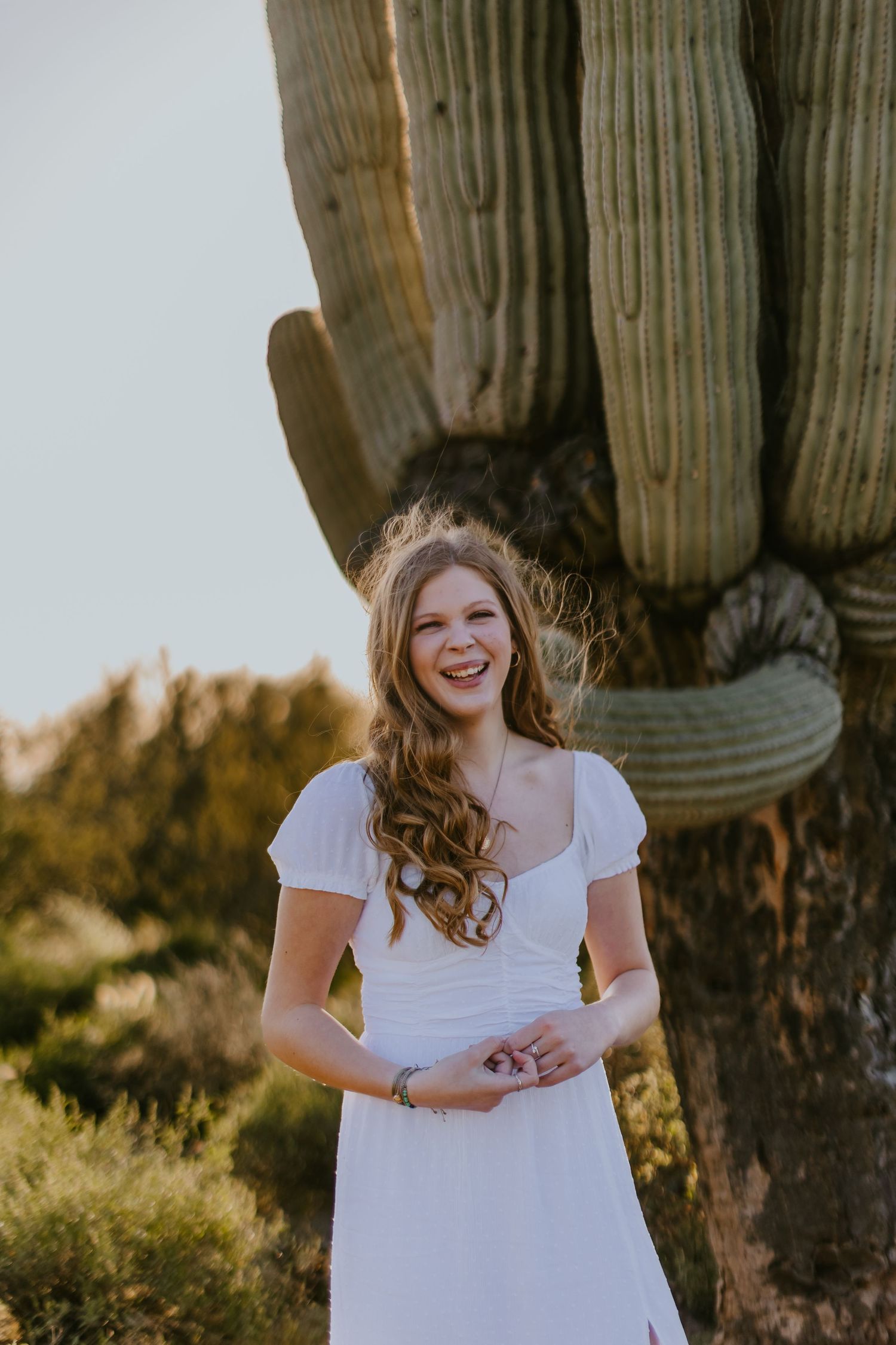Joyful portrait in white dress next to towering saguaro cactus at golden hour.