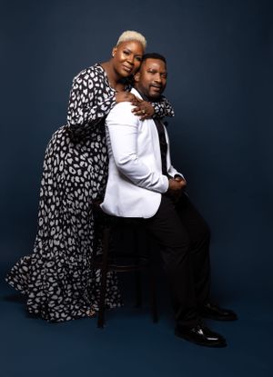 Elegant studio portrait with couple in white blazer and flowing printed dress seated on chair.
