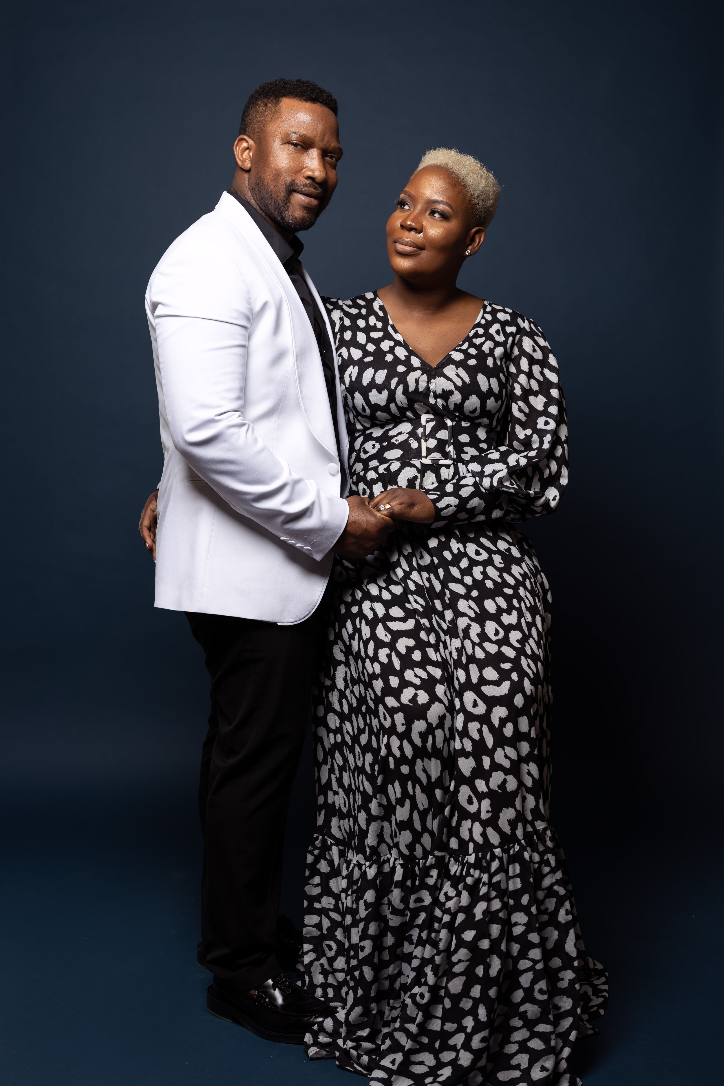 Stylish couple poses in white blazer and flowing printed dress against dark studio background.
