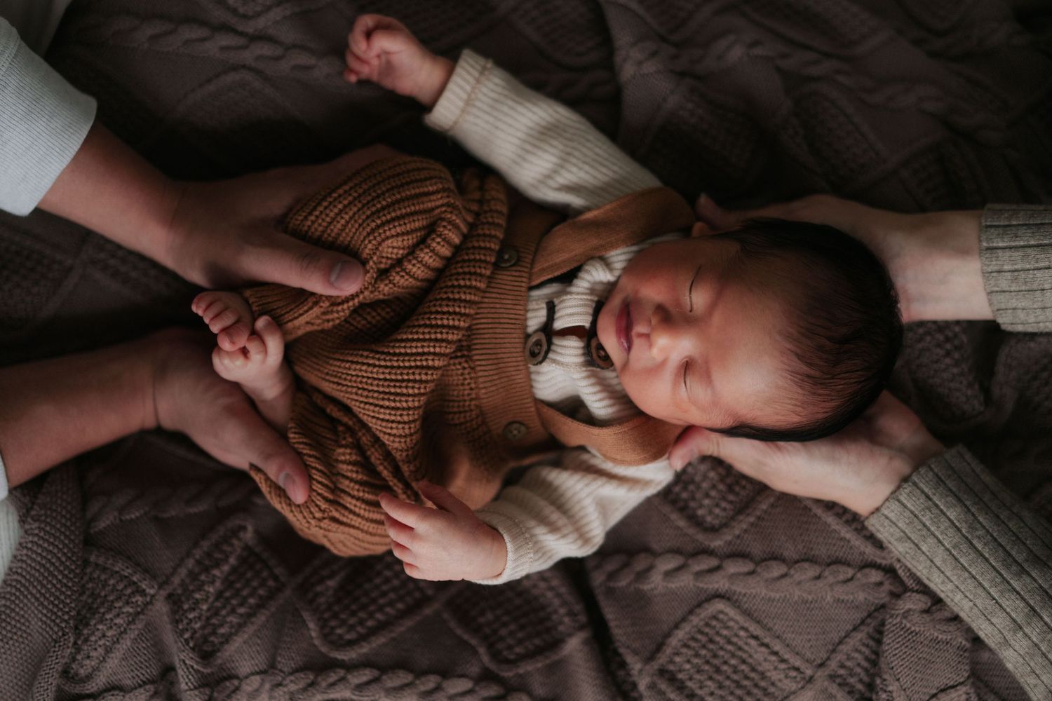 Baby in brown sweater lies peacefully on a quilted blanket.
