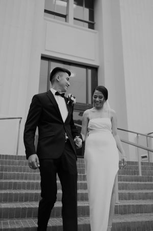 A black and white photo of a couple in wedding attire standing on steps outside a building.