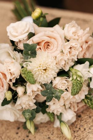 Elegant wedding bouquet featuring blush roses, white dahlias, and green foliage on textured surface.