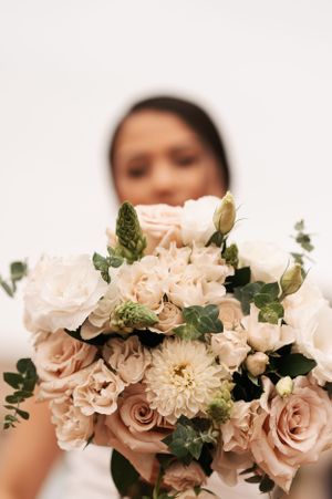 Close-up of a blush pink and white wedding bouquet with roses, dahlias, and greenery against a light background.