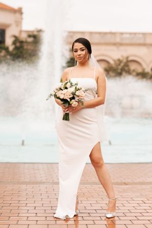 Bride in elegant white dress poses with bouquet near fountain.