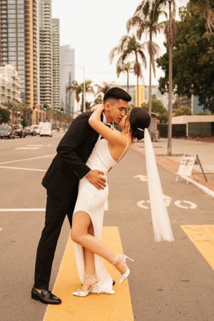 A romantic moment between a couple in formal attire on a city street with palm trees and buildings.