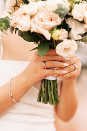 Close-up of hands holding a romantic blush pink wedding bouquet with delicate bracelet and ring details.
