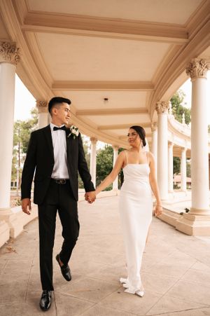 Elegant couple walking hand in hand under ornate colonnade in wedding attire.