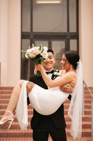 Groom in black tuxedo carrying bride in white dress with floral bouquet on brick steps.