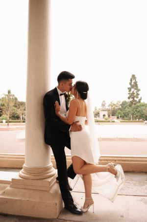 Playful wedding pose with bride lifting leg while embracing against a columned architecture.