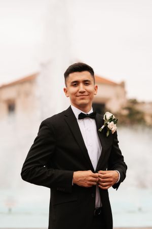 Groom in classic tuxedo with boutonniere poses against light background.