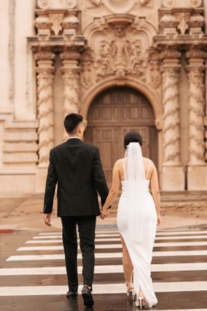 Couple in wedding attire walking towards ornate baroque-style church entrance.