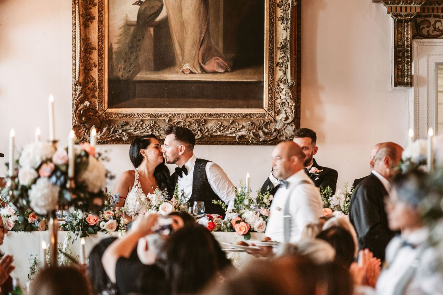 Wedding reception guests dining at an elegant venue with ornate gold-framed artwork on the wall.