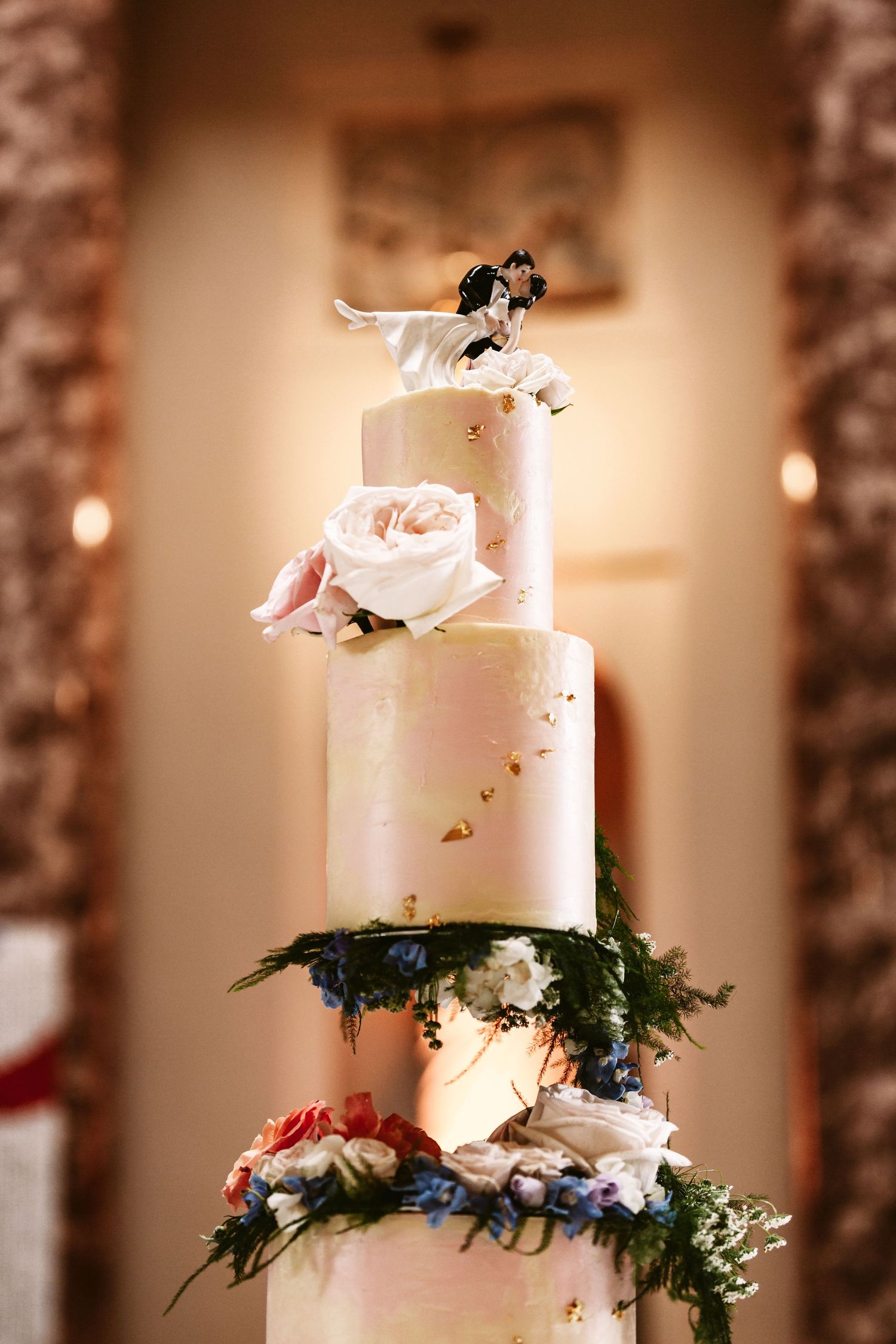 Elegant white wedding cake decorated with roses and small figurine topper against warm lighting.