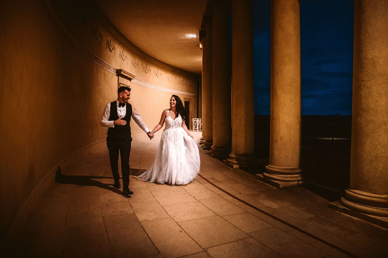 A couple walks hand in hand through an elegant colonnade at dusk.