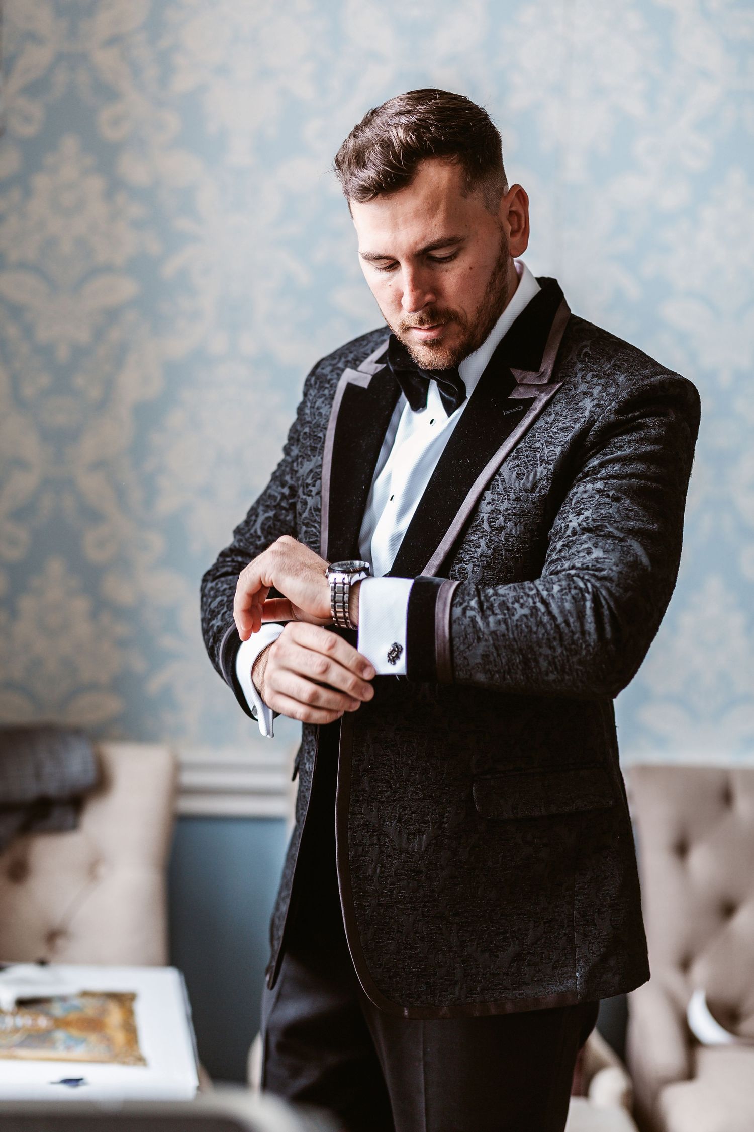 A person adjusts their cufflink while wearing a textured dark formal jacket with bow tie.