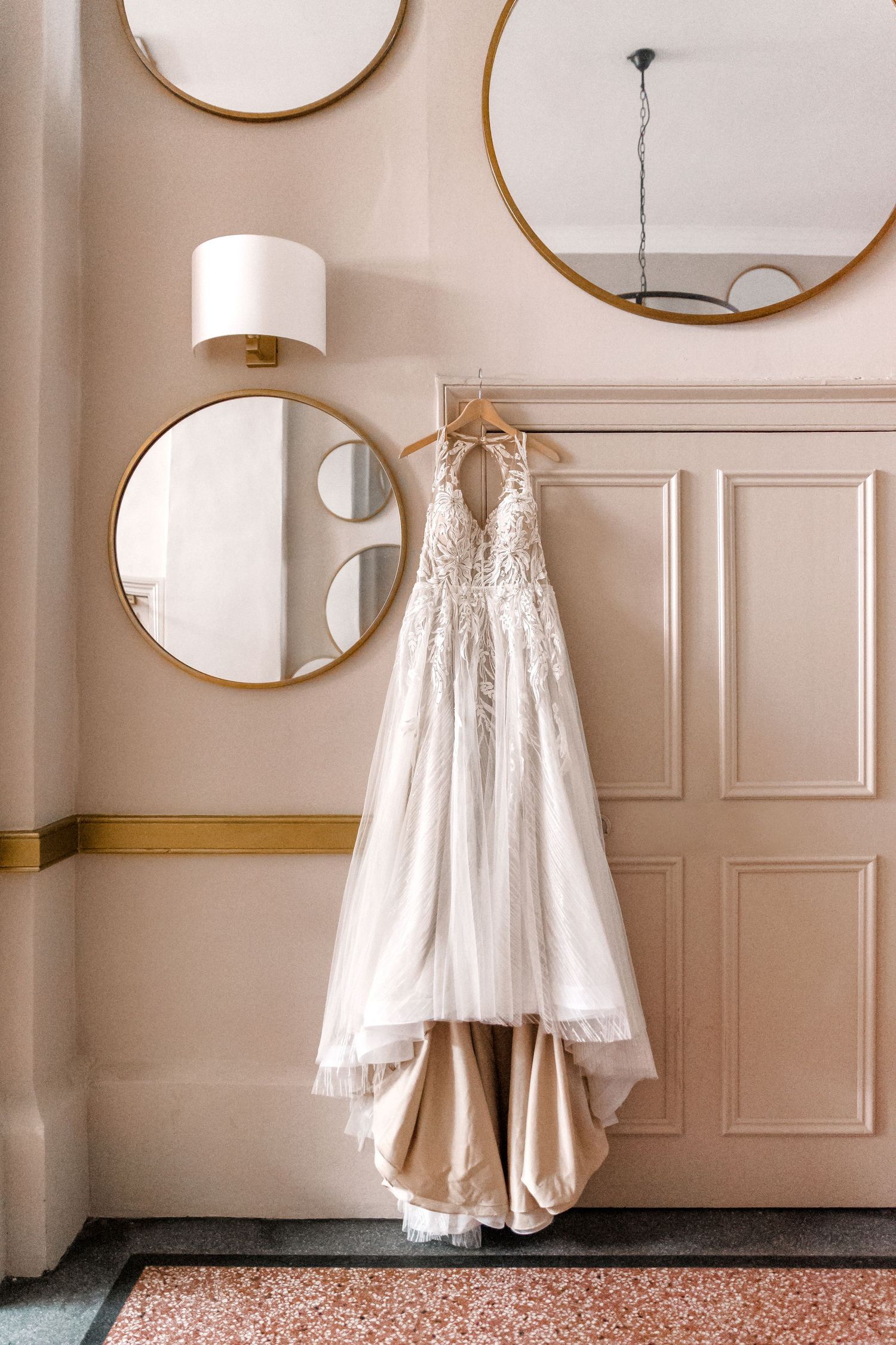A white lace wedding dress hanging on a wall with three round gold-framed mirrors.