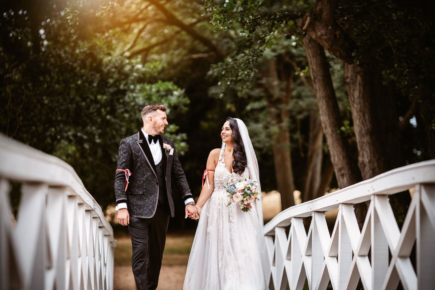 Couple walks along white latticed bridge during sunset wedding photography session.