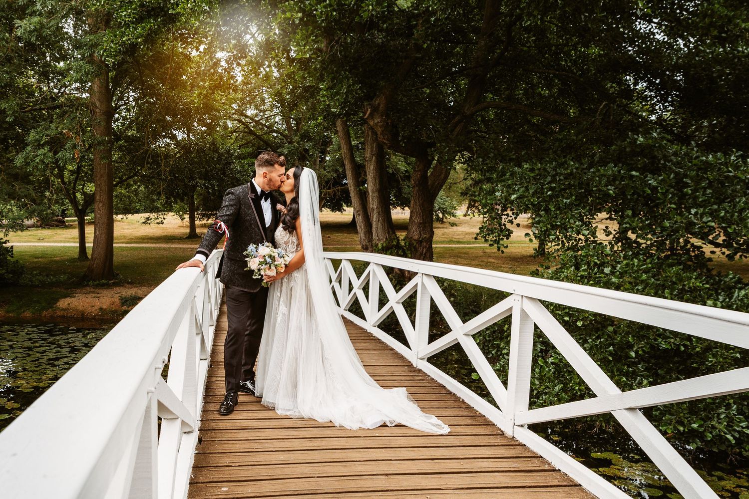 Couple in formal attire share romantic moment on white wooden bridge surrounded by trees.