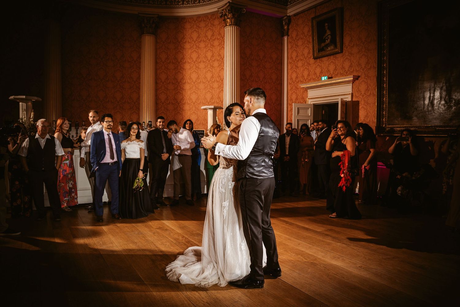 First dance at wedding reception in dimly lit historic ballroom with watching guests.