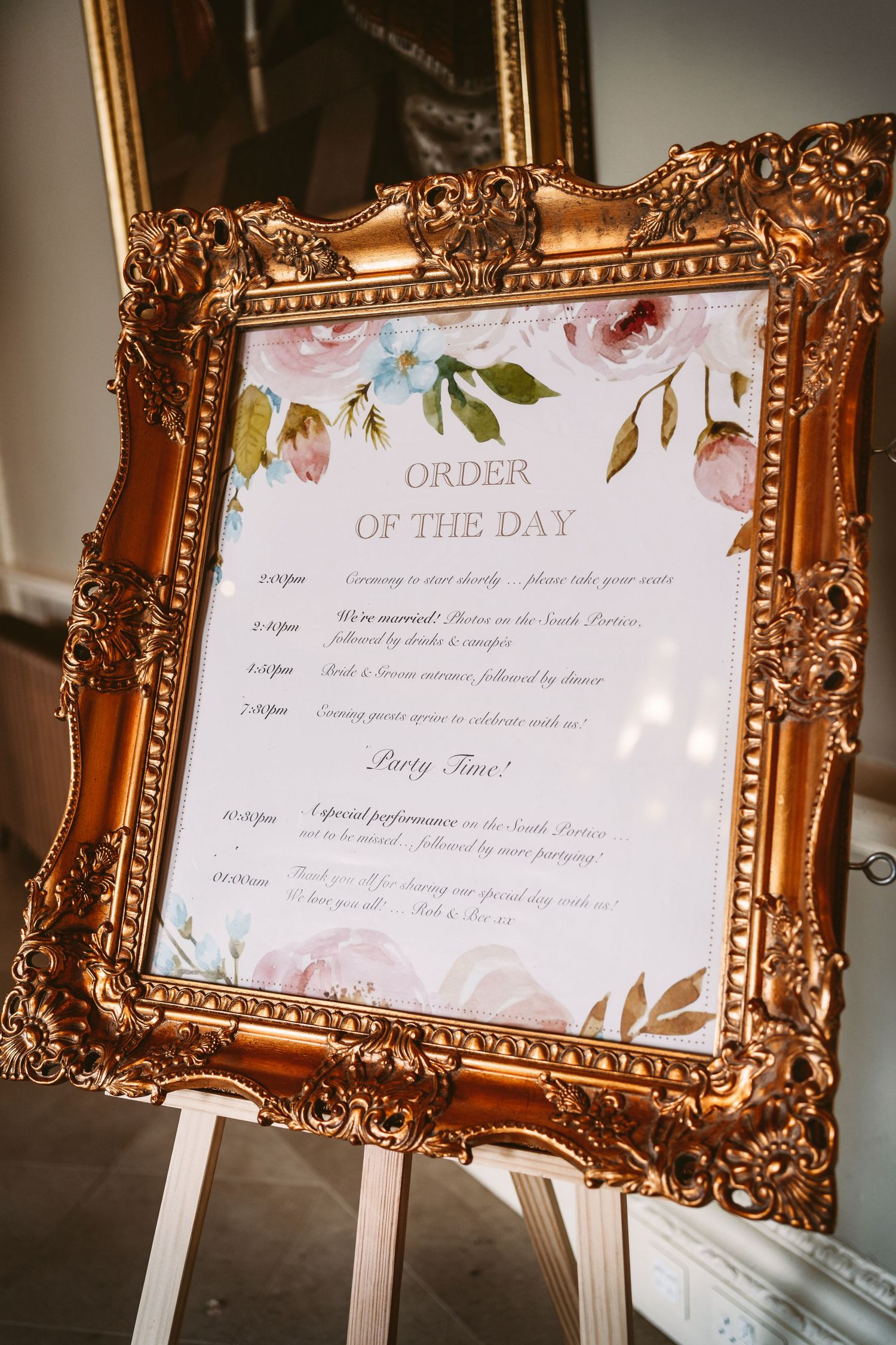 Ornate gold frame displaying wedding day timeline schedule with floral design on white paper.