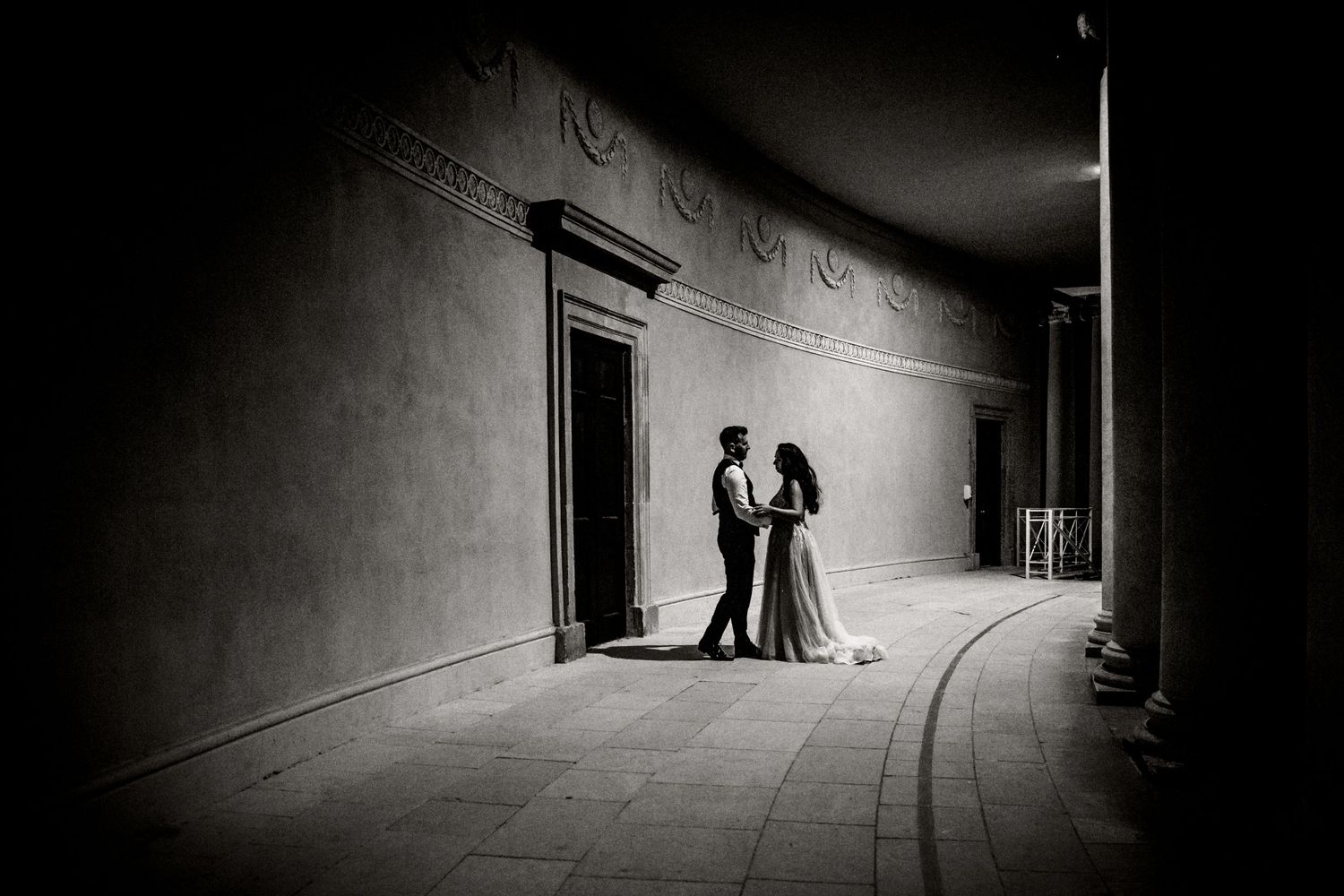 A black and white photograph capturing a couple in a dramatically lit curved corridor.