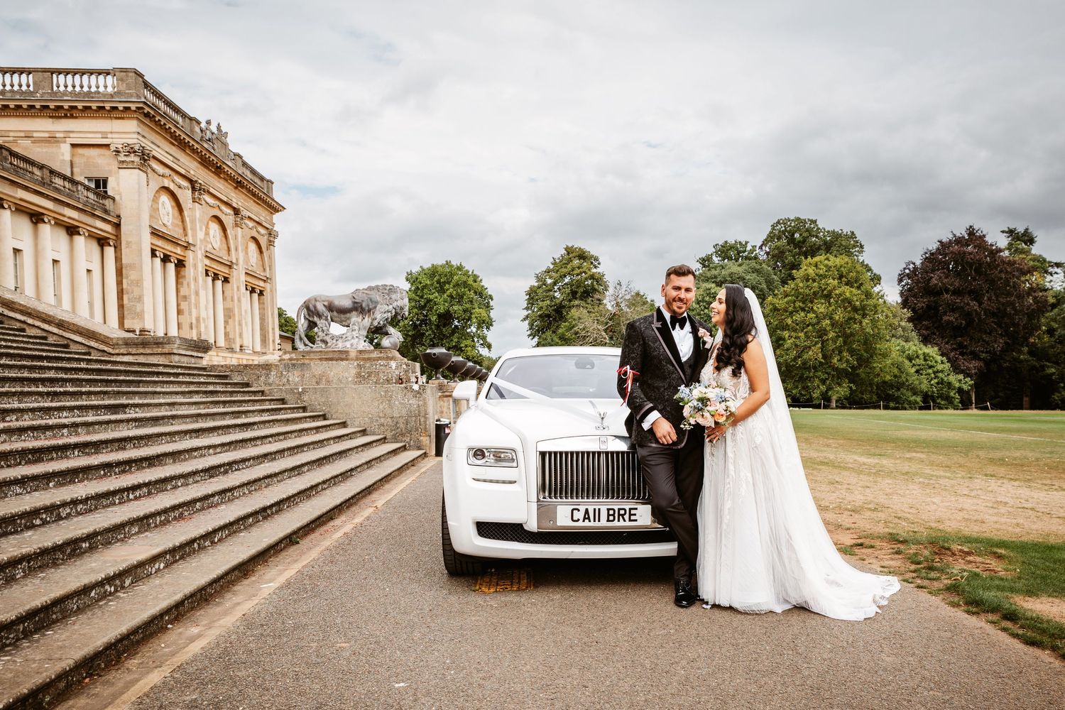 Wedding couple poses with white Rolls Royce in front of grand stone mansion.