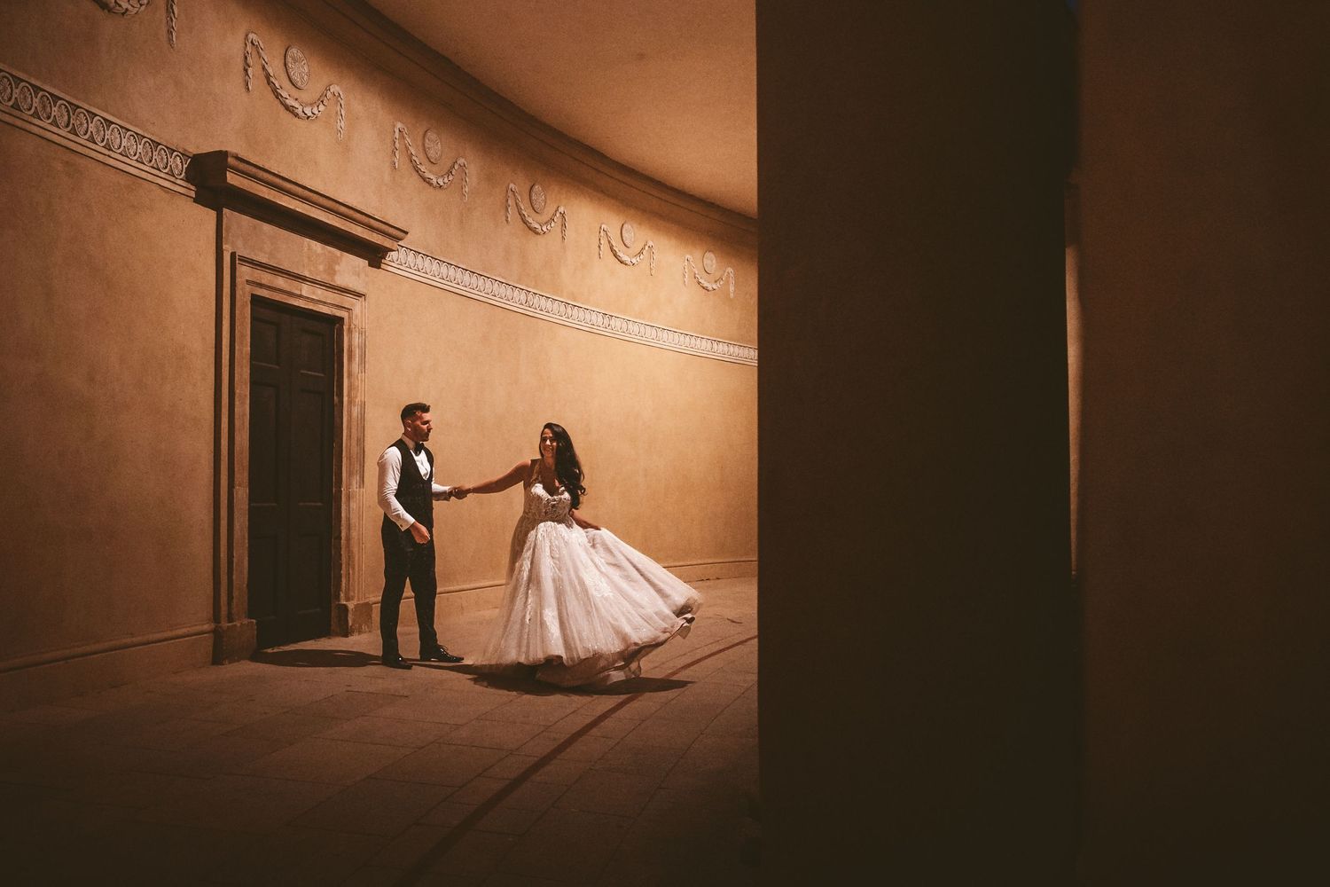 A couple dances together in front of a wooden door in a curved corridor with decorative molding.