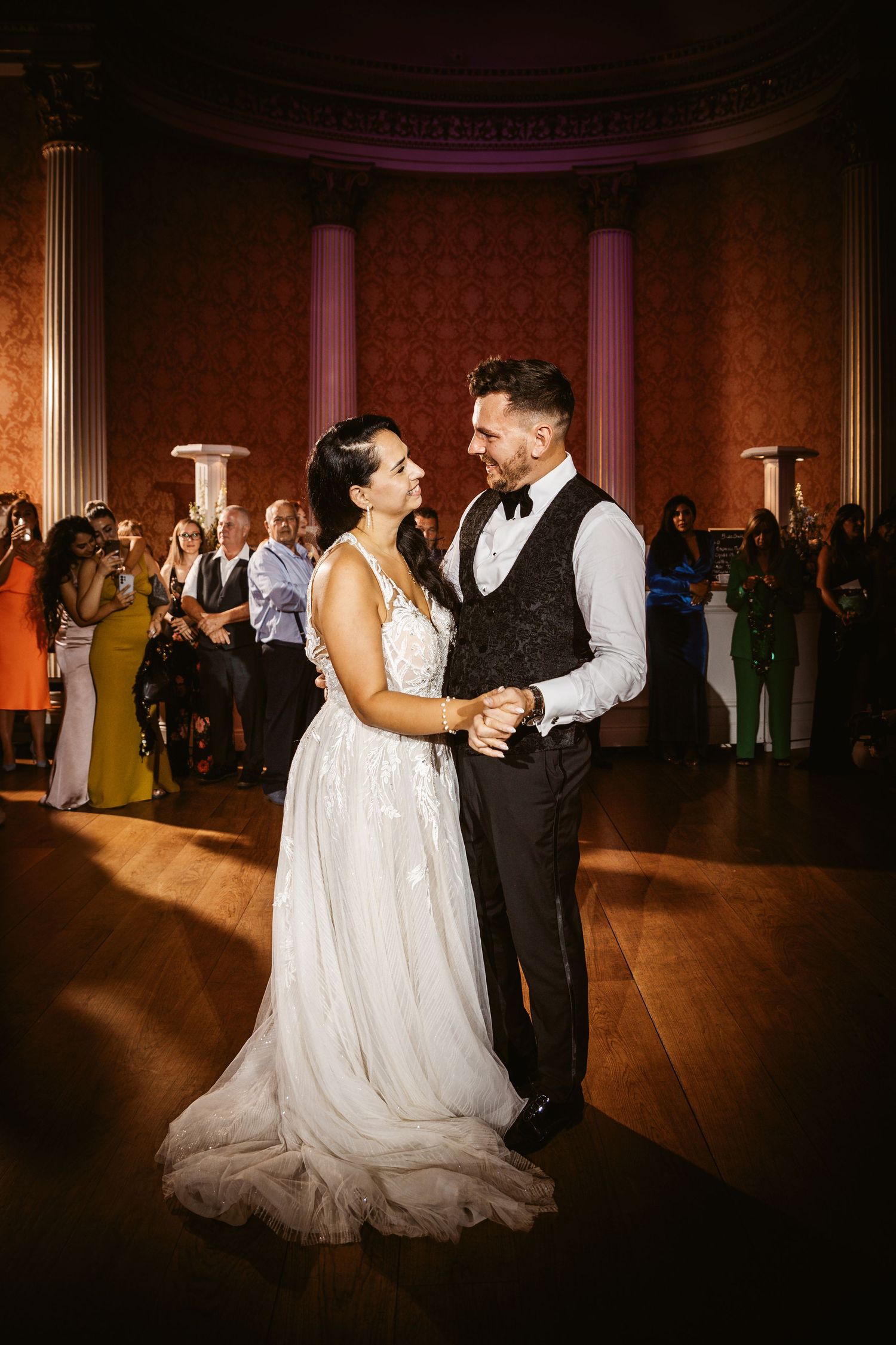Newly married couple dances intimately in a grand ballroom with classical architecture and columns.