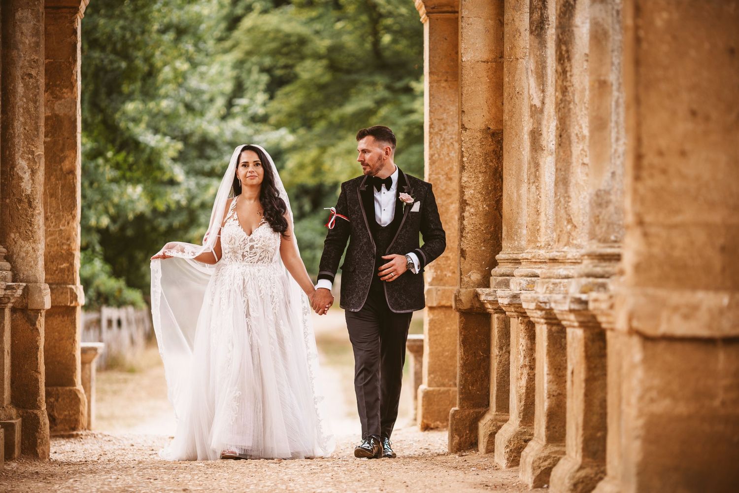 Couple poses between ornate stone columns during elegant outdoor wedding shoot.