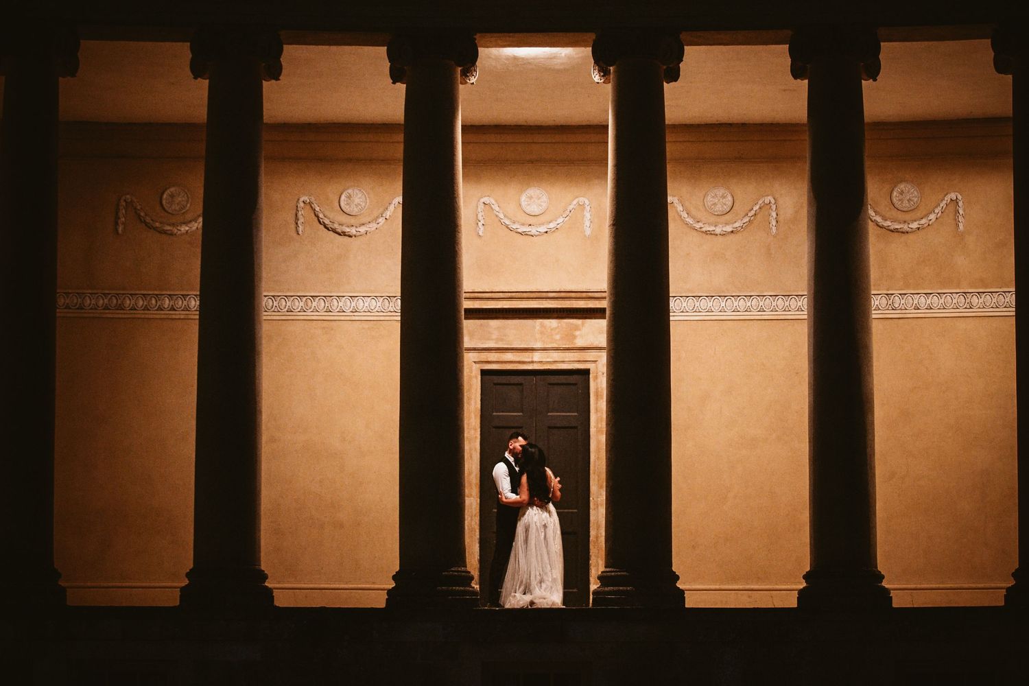 A romantic moment captured between classical columns at night with warm lighting.