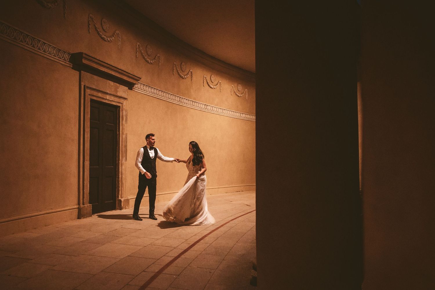 A couple shares an intimate dance in a warmly lit curved hallway with ornate architectural details.
