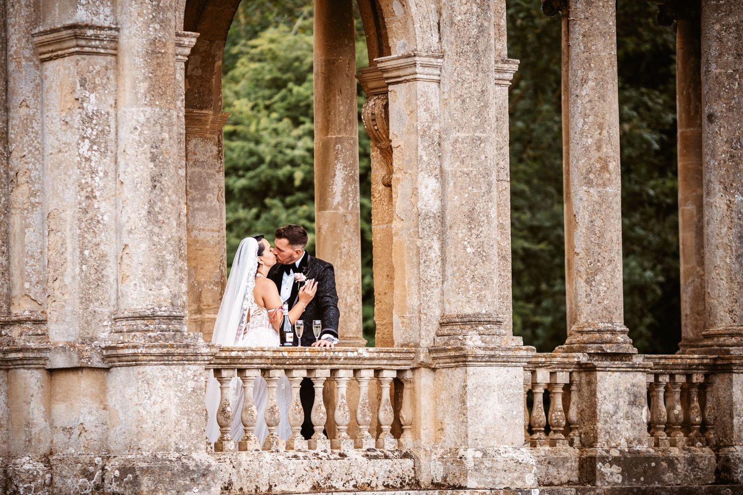 Couple shares intimate moment between classical stone columns during outdoor wedding shoot.